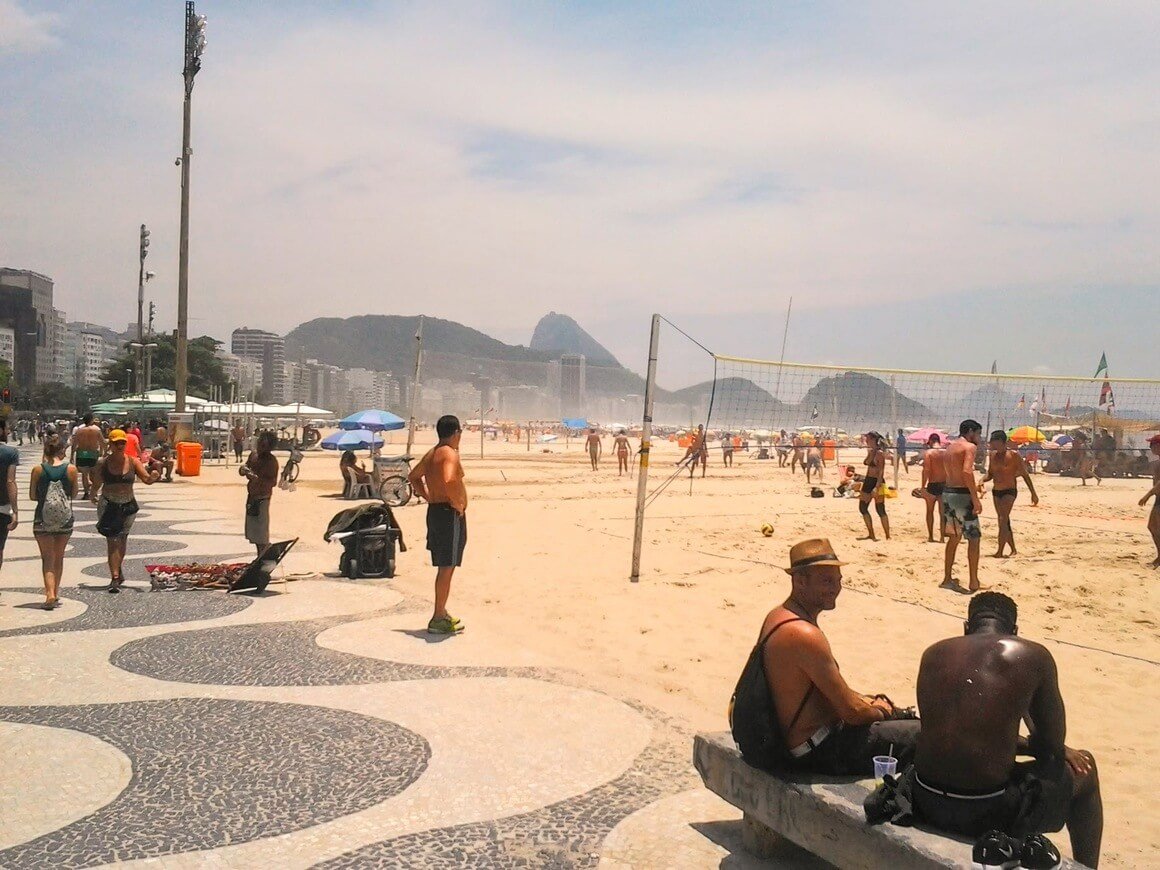 People walking, sitting and playing volleyball on a sunny day at Copacabana Beach in Rio de Janeiro.