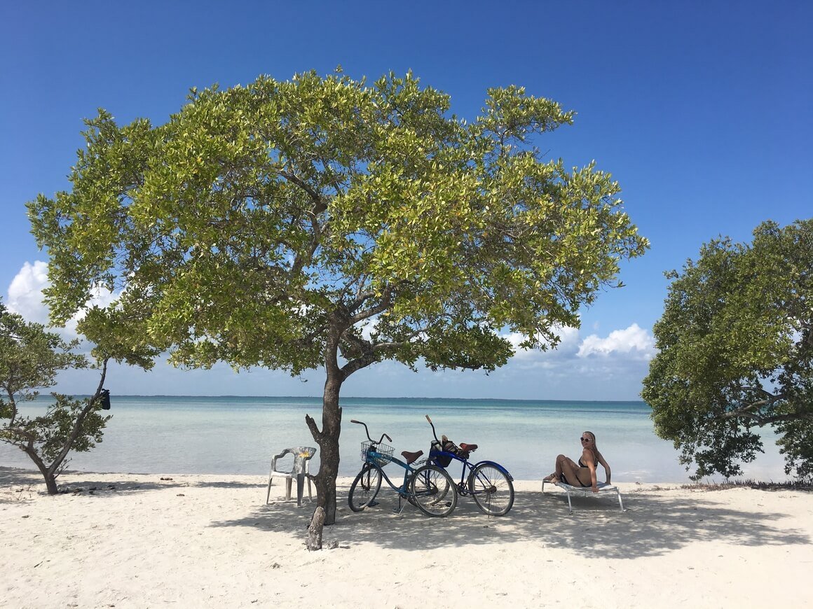 Laura lying on a sun lounger under a tree text to two bikes on white sand next to a blue  Caribbean sea in Holbox, Mexico