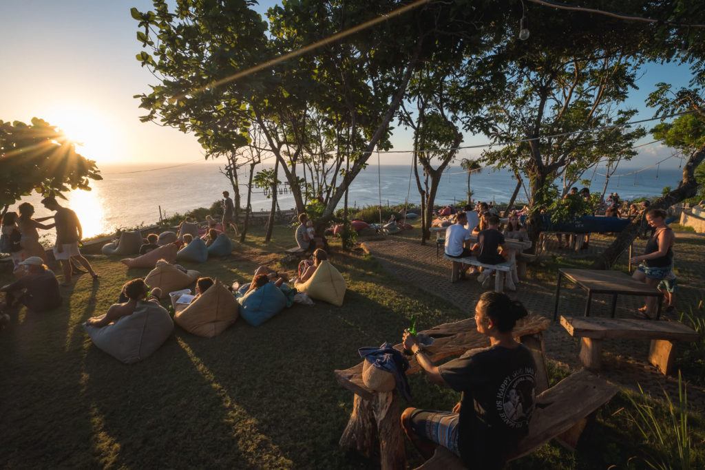 A group of people on bean bags at a bar or hostel are sitting on a cliff watching the sunset in Uluwatu, Bali