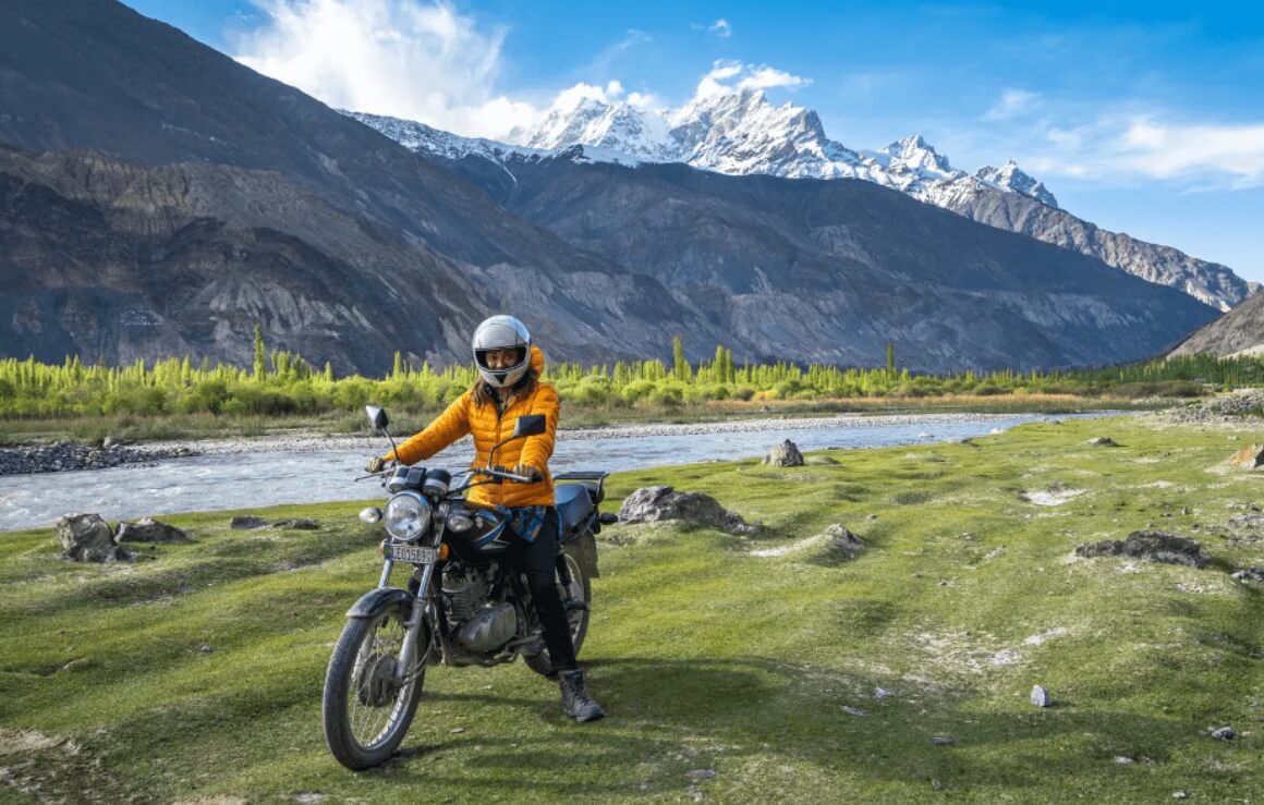 girl in a yellow jacket riding a motorcycle in a lush green meadow with snowcapped peaks in the background