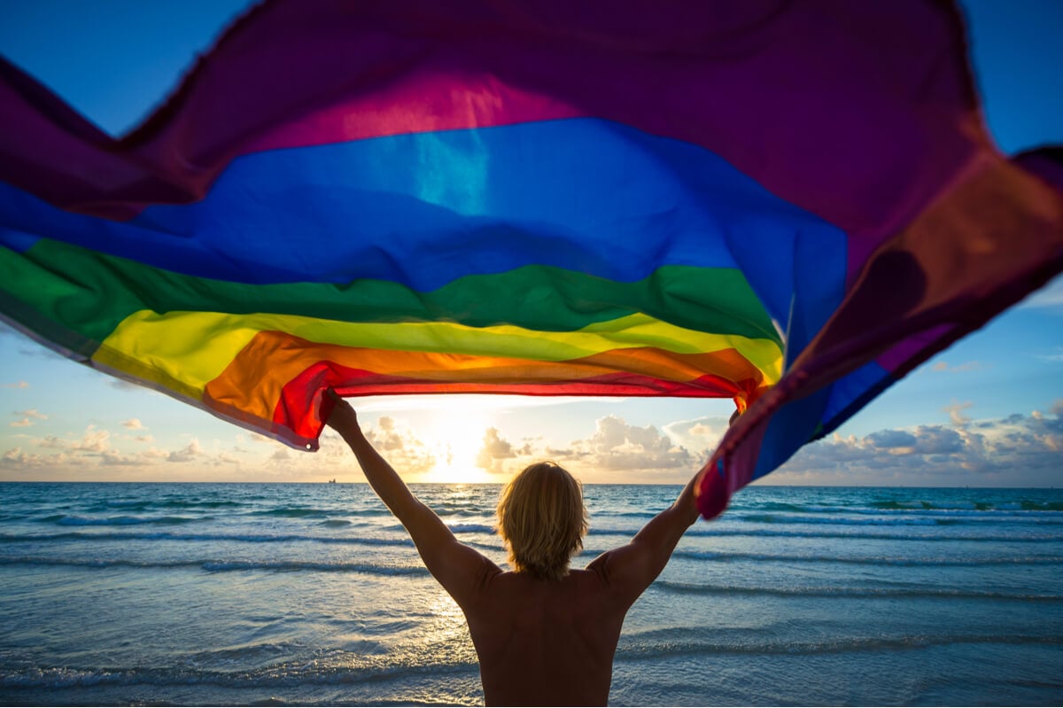 A man holding a rainbow flag on the beach celebrates his LGBT travel life.