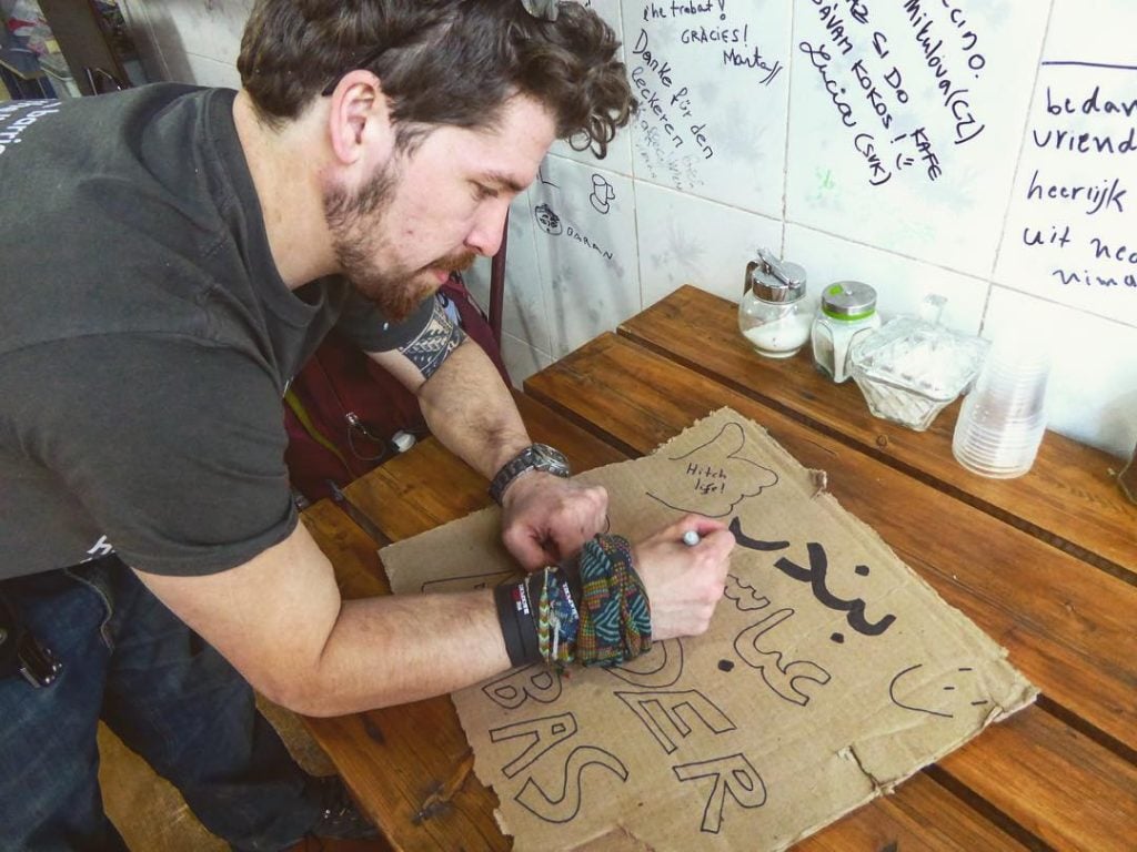 man making a cardboard sign to overland travel by hitchhiking