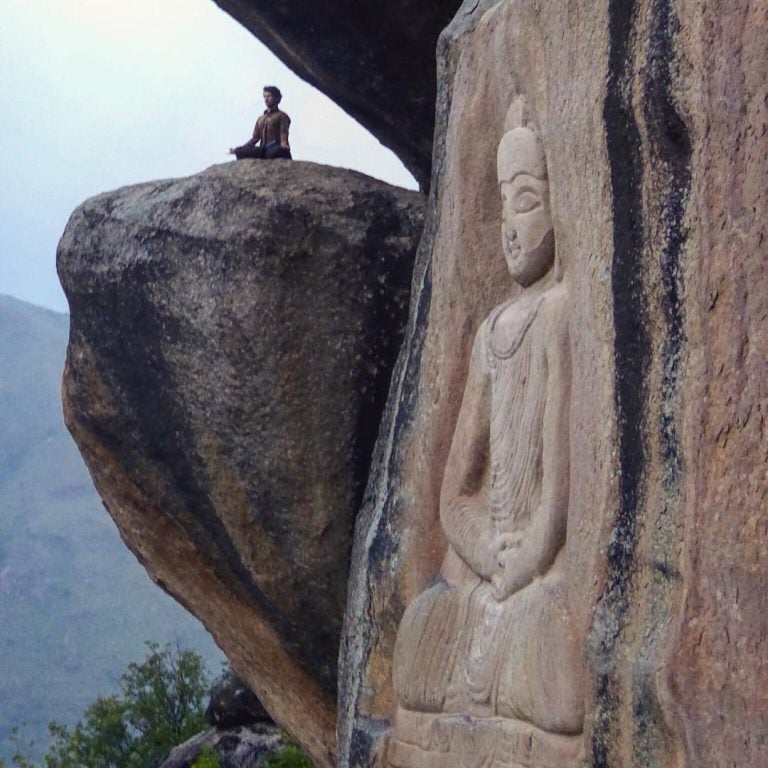 A man sitting in the lotus yoga position sits on a rock at the end of a cliff with a buddha carving in the cliff in the foreground