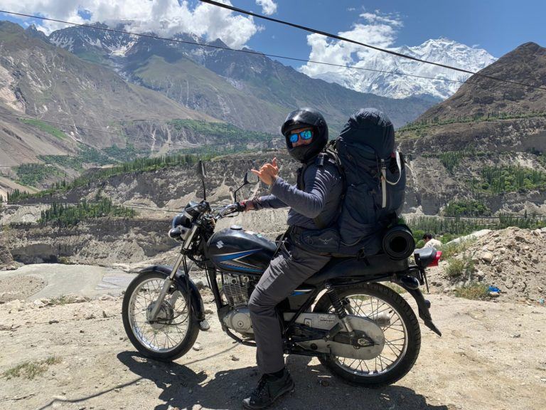 A man on a motorcycle/ motorbike with a large backpack with camping gear attached doing a shaka sign whilst stopping for a break whilst riding through the mountains. In the background is a valley with several mountains, some covered in snow