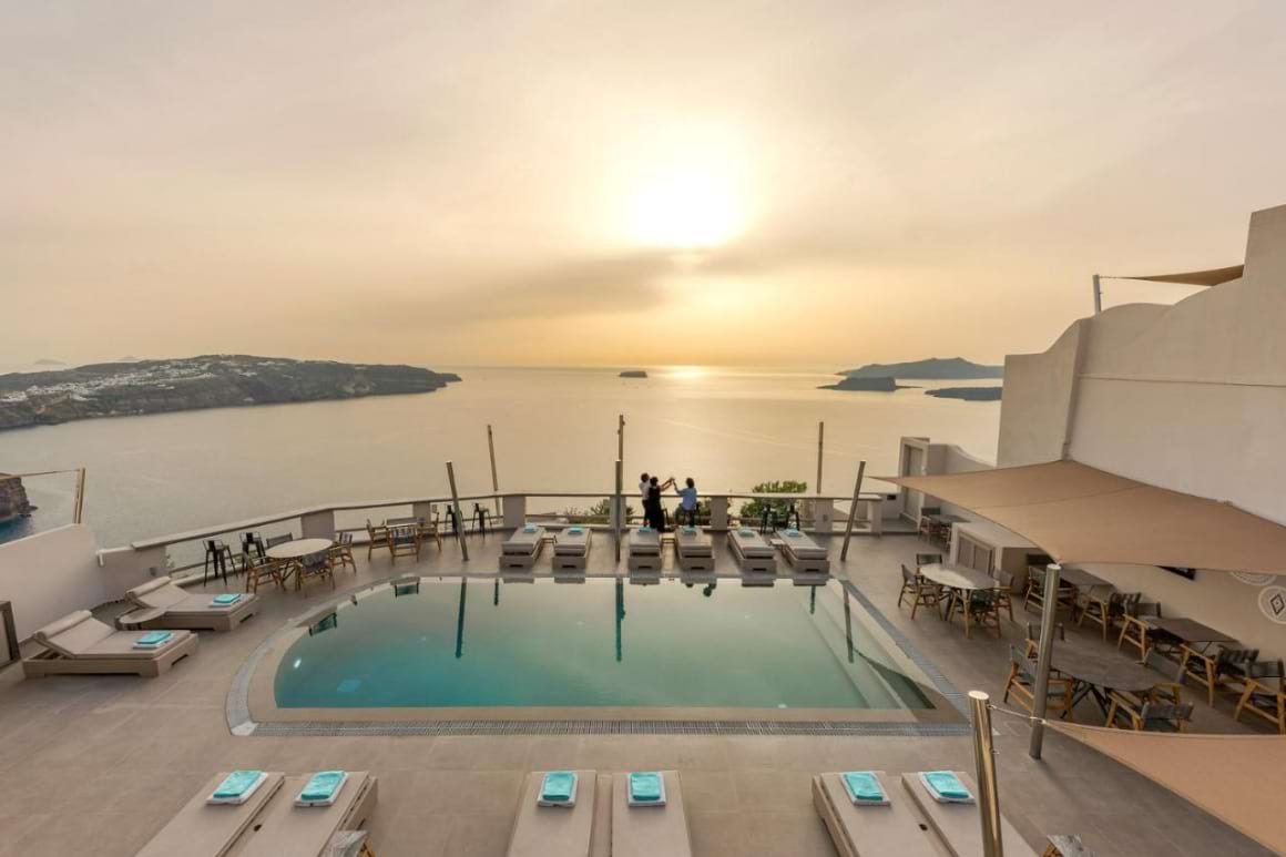 A swimming pool sits on the rooftop of a Santorini hotel with the ocean and a colorful sunset sky in the background