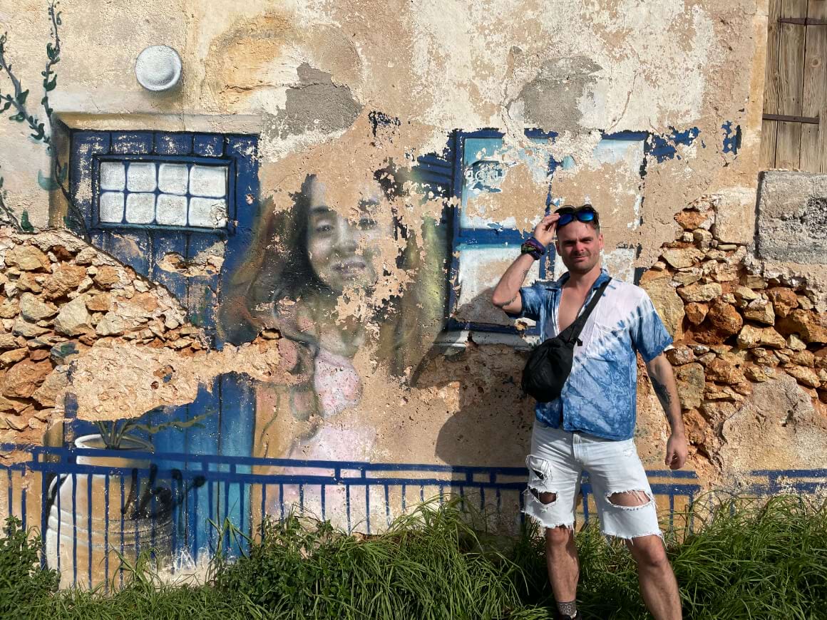 A man standing by a wall with street art wearing a sling. 