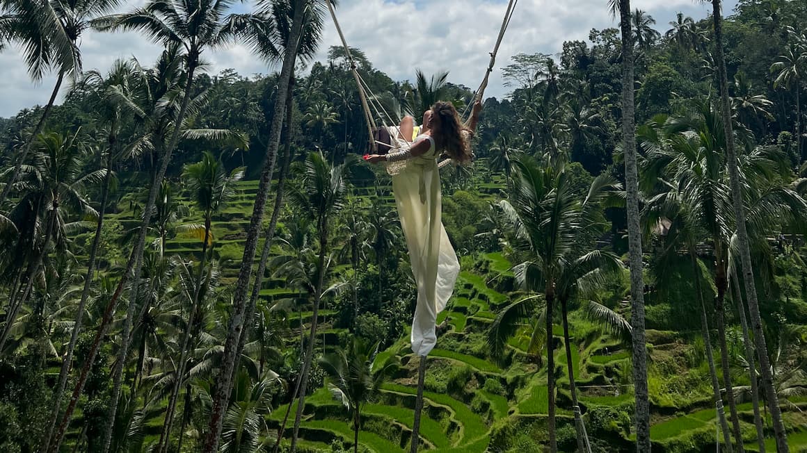 A woman in a white dress swinging on a large swing over lush, green rice terraces and palm trees in a tropical landscape.