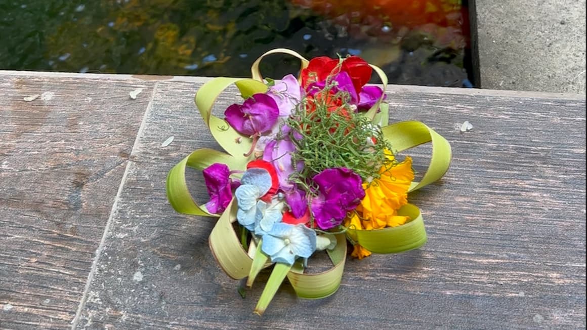 A colorful arrangement of flowers in a woven basket made of green leaves placed on a wooden surface, with a blurry water background.