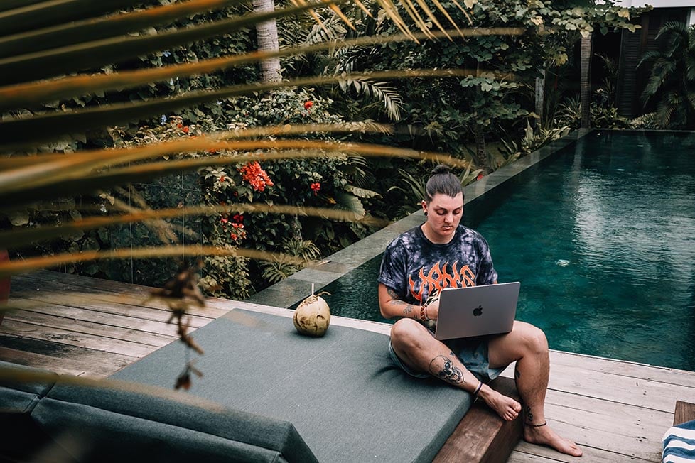 Nic Hilditch-Short sitting on a poolside lounger with a laptop on their lap, surrounded by lush tropical foliage and a fresh coconut beside them, with natural wooden decking leading to a serene swimming pool