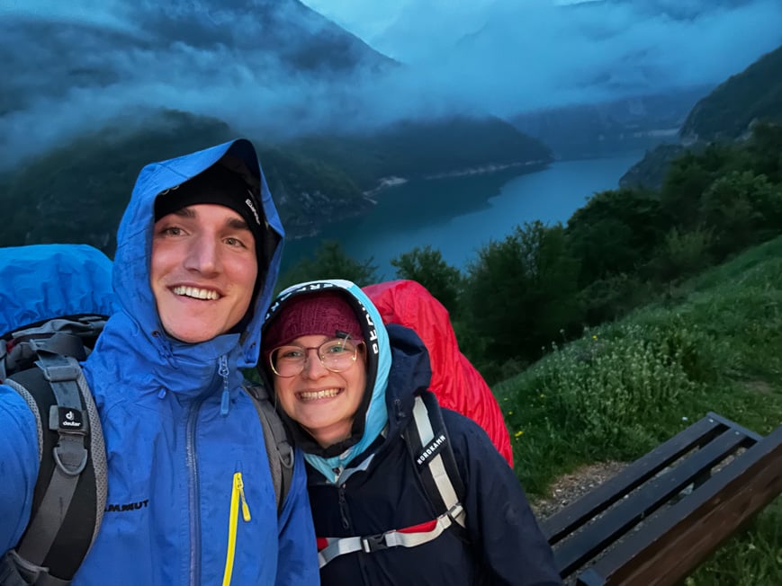 a couple wearing raincoats and carrying big backpacks hiking in a forest above a lake