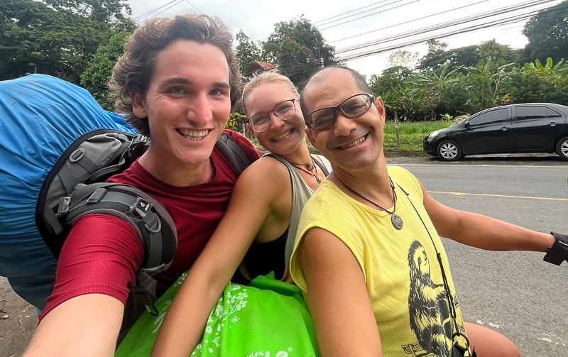 a couple hitchhiking on a motorbike with the guy carrying a huge blue backpack