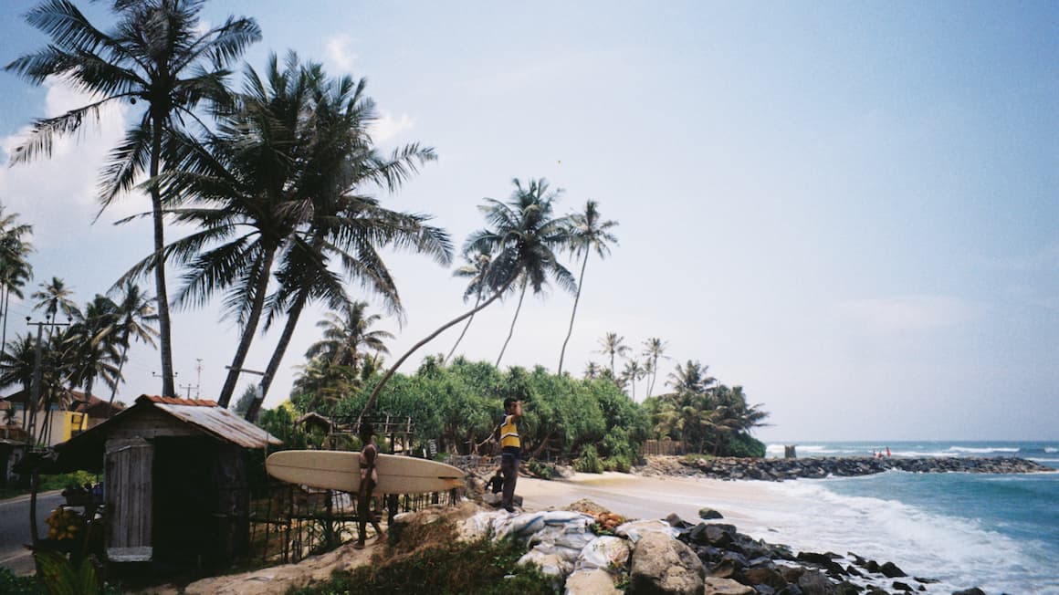 a surfer walking over rocks towards the ocean at a beach with palm trees in Sri Lanka 