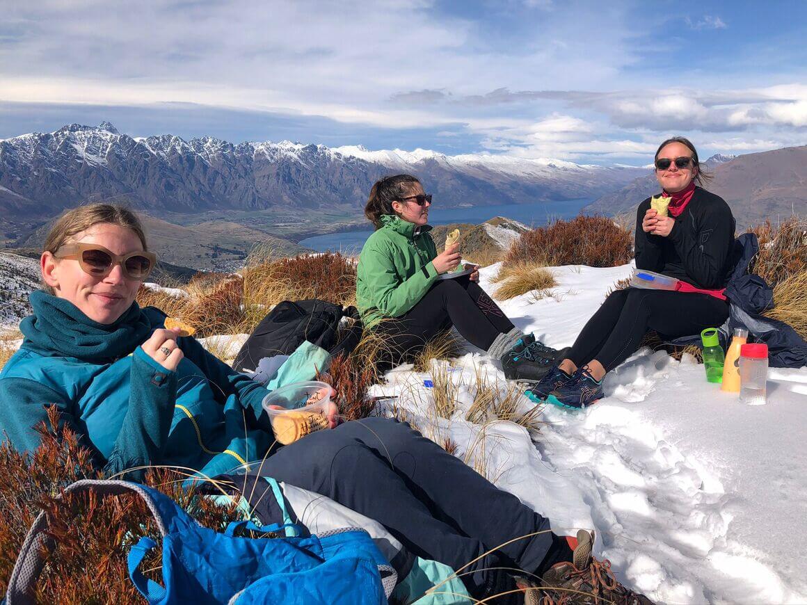 Group of friends enjoying snacks on top of an icy summit, surrounded by mountain views.