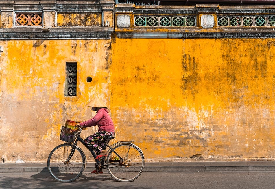 A woman on a bike with a traditional hat on rides passed the yellow walls of Hoi An, Vietnam