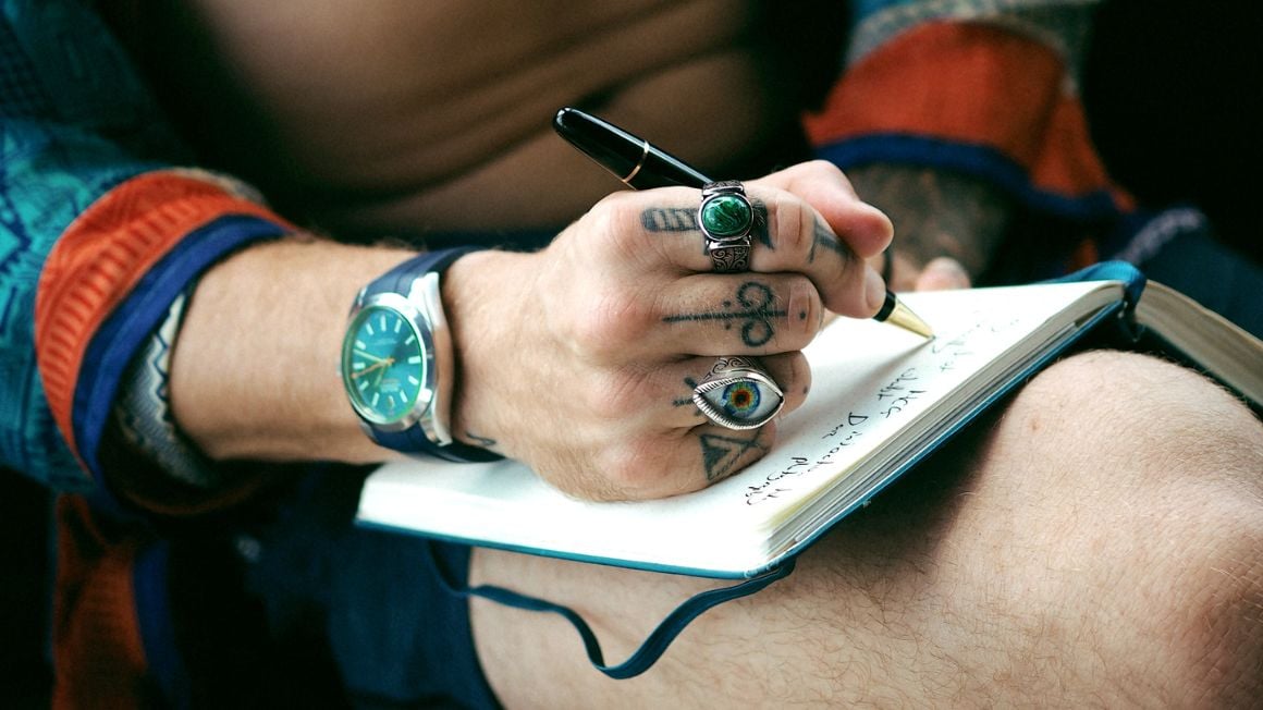 close up of man's hand with rings and tattoos writing on a journal