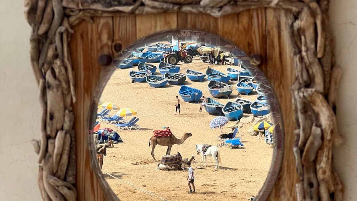 a mirror showing a reflection of the beach, with blue boats docked on the shore and a camel standing on the sand in taghazout morocco