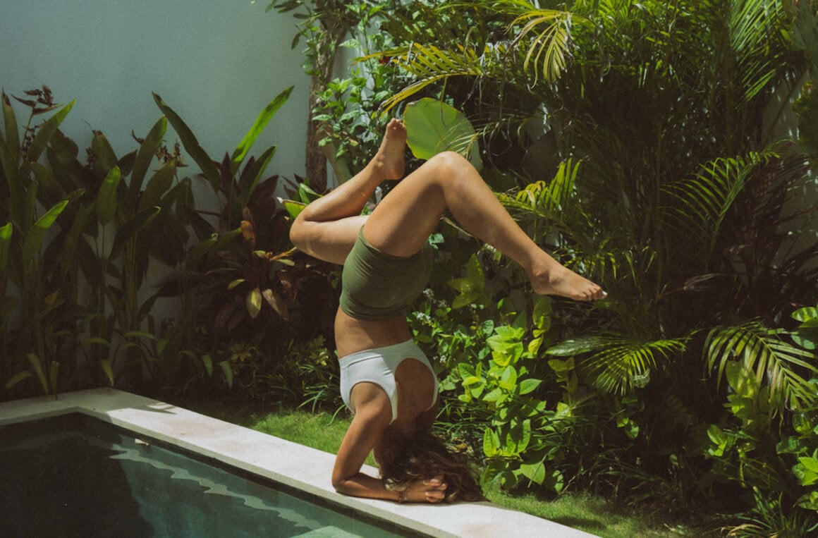 A woman (Amanda) performs a handstand yoga pose beside a pool in a vibrant garden setting.