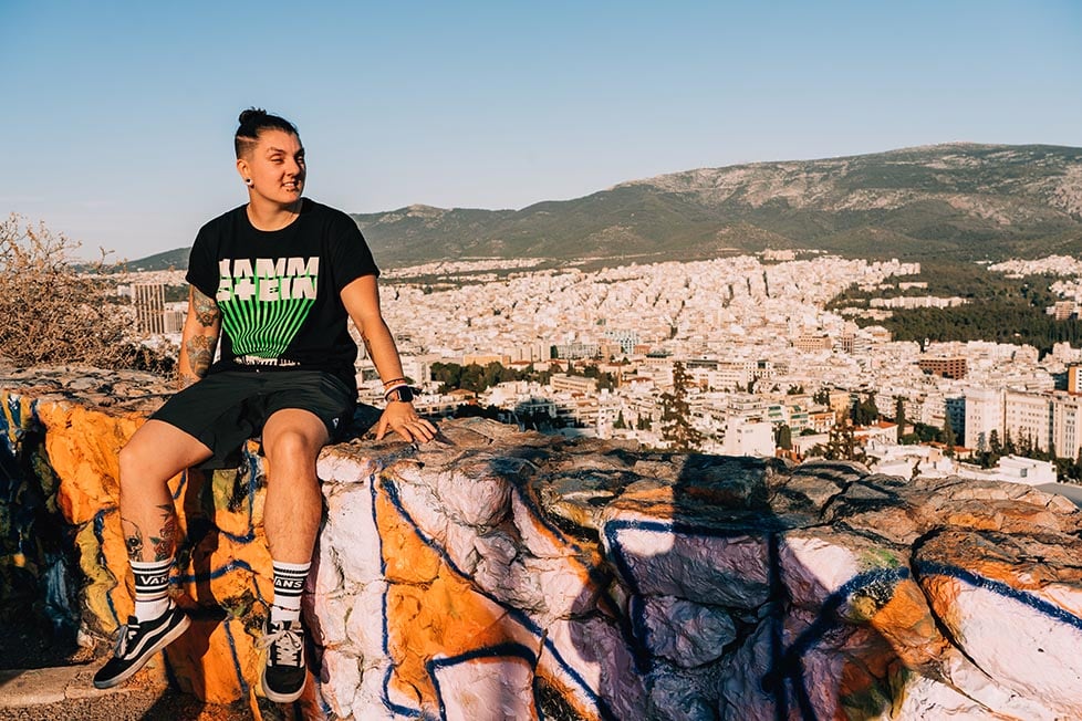 Nic sitting on a wall covered in graffiti overlooking Athens, Greece below and a mountain behind it. They have extra ordinarily muscular calves!
