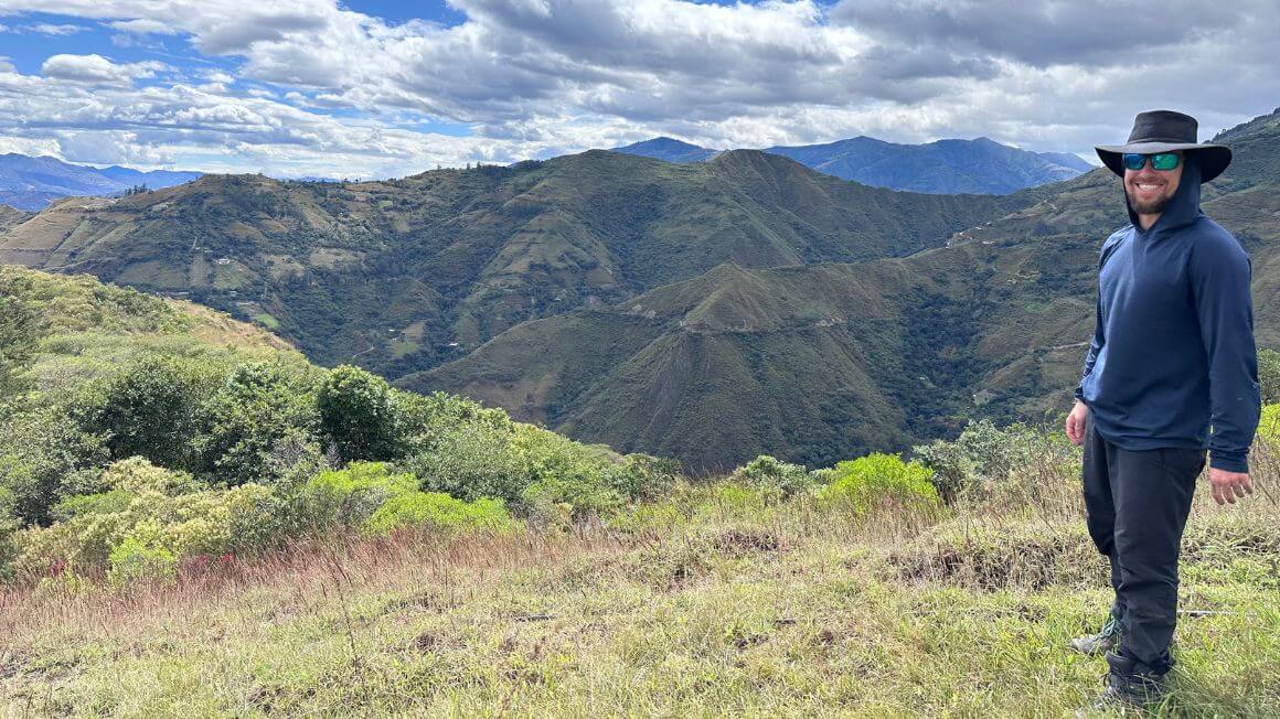 Will standing on a hillside with lush jungle covered mountains behind him whilst also wearing a cowboy hat in Ecuador