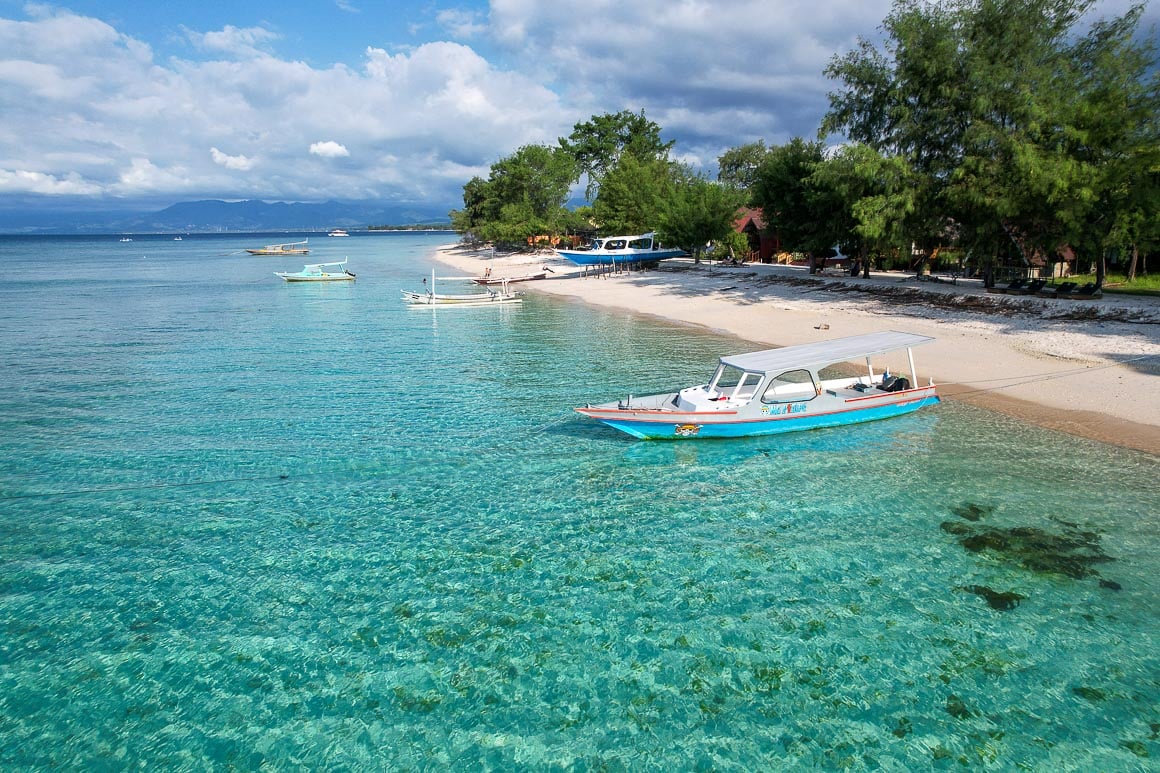 a small boat floating on bright blue turquoise water with the beach and large green trees behind it in gili meno in indonesia on a sunny day