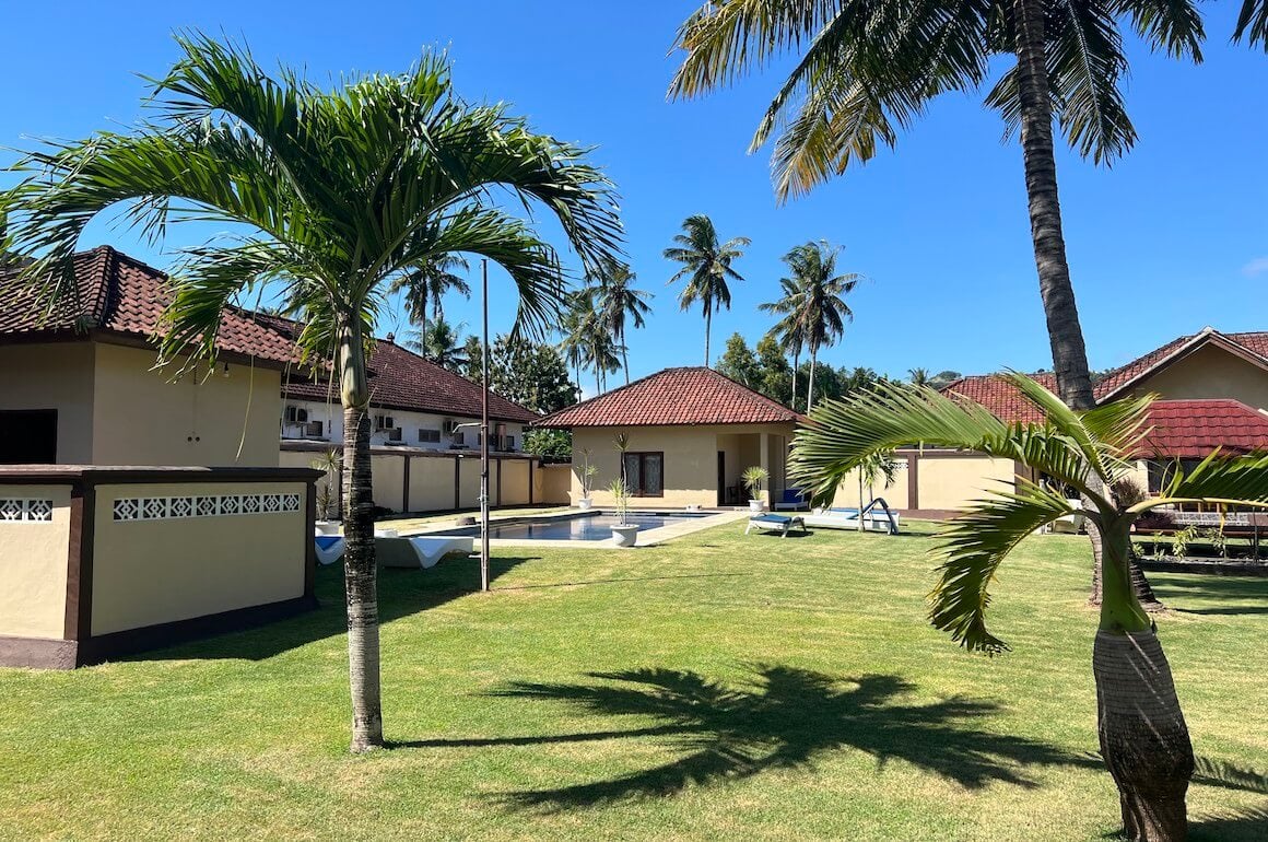 a clear blue sky day shot of the yard of a lombok homestay with multiple palm trees green grass and one of several pools