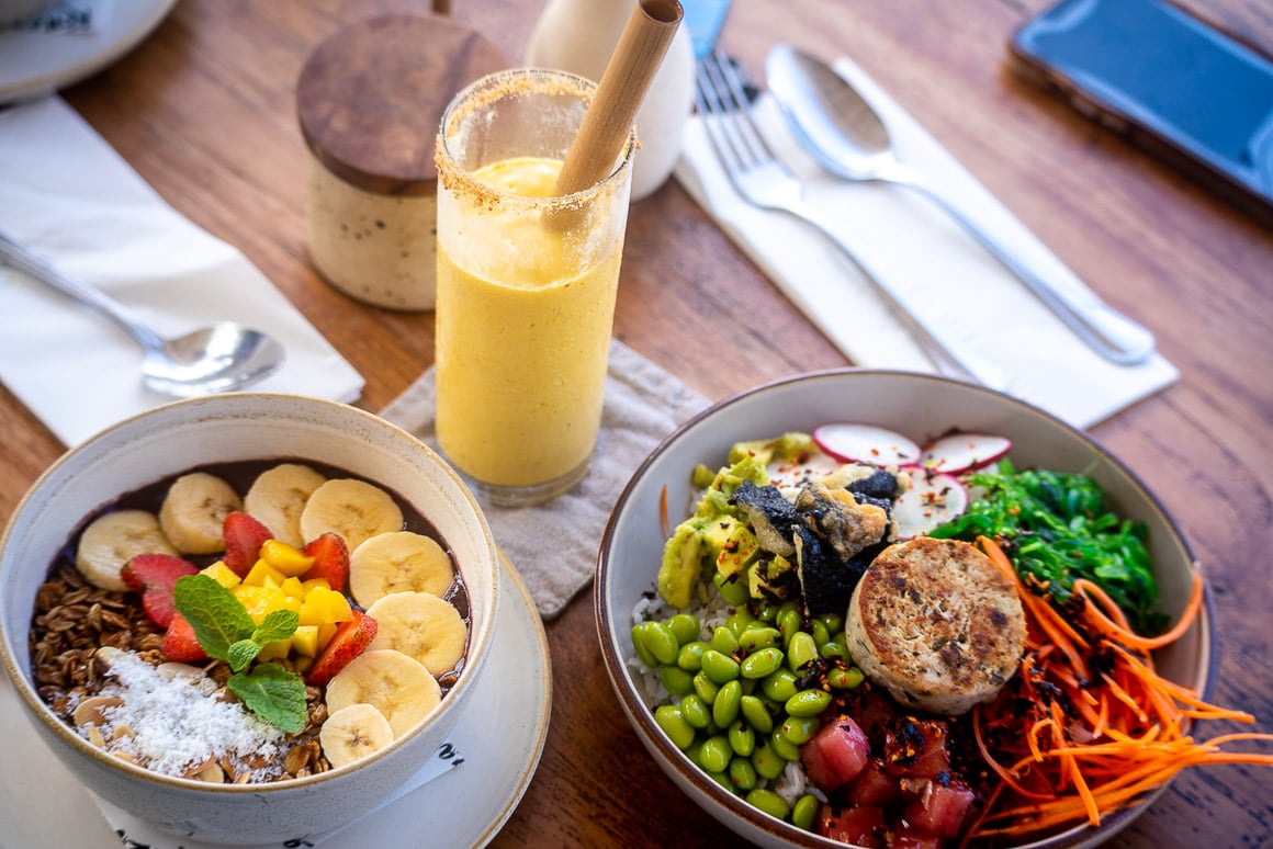 a fruit filled smoothie bowl next to a colorful poke bowl on a wooden cafe table in kuta lombok