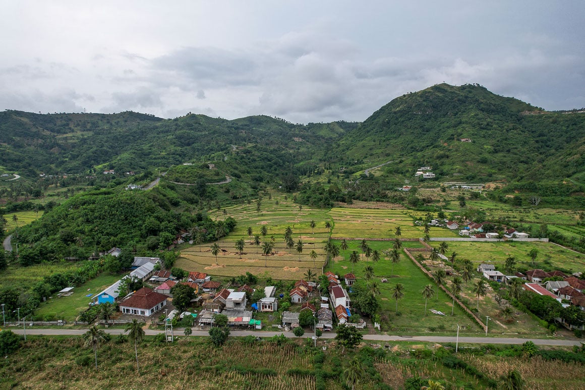 a lush green drone shot of a village area with numerous hills and rice fields with small houses with reddish orange roofs in lombok indonesia