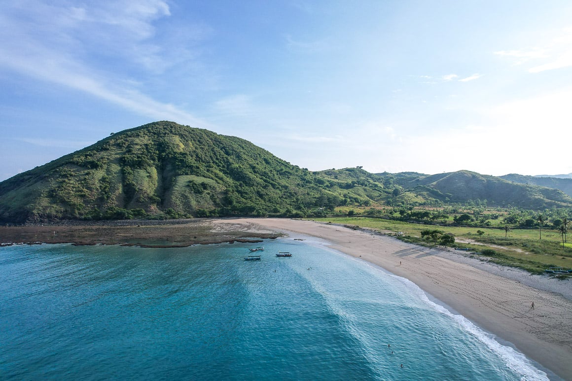 drone view of bright blue calm sea water crashing into a tan beach with a huge green forested hill in the background in lombok