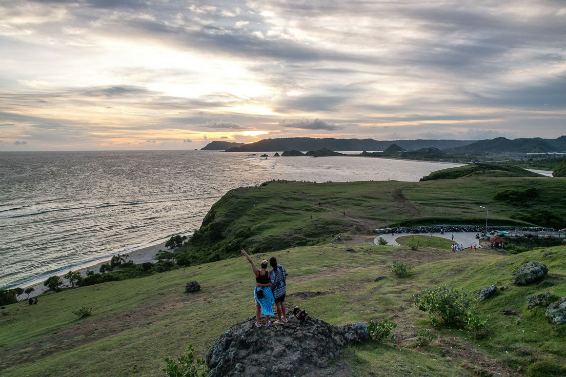 a couple standing on rock on the green bukit merese hill facing the sea and a grey yellow sunset in lombok in indonesia