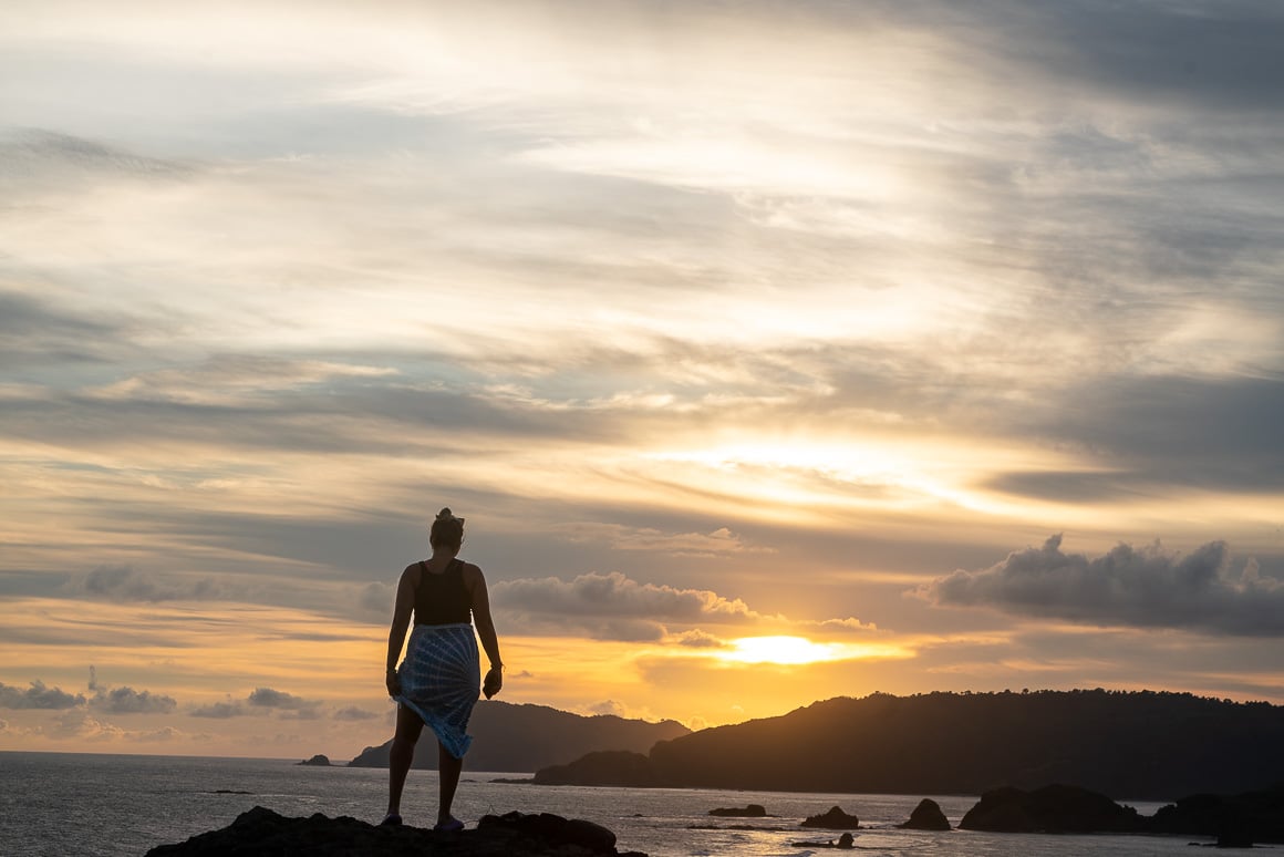 a girl standing on a rock wearing a blue and white sarong watching an orange sunset over the seas and cliffs of kuta lombok