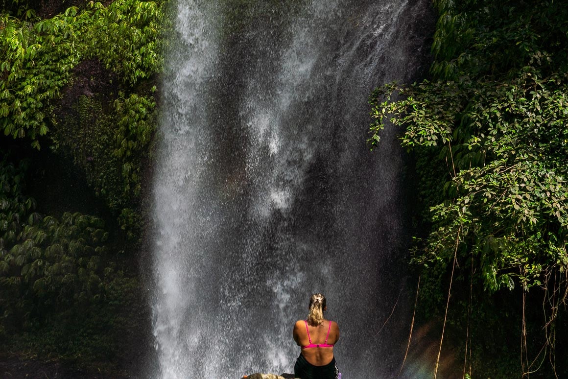 a girl sitting and facing a huge waterfall coming down in front of her in north lombok in indonesia