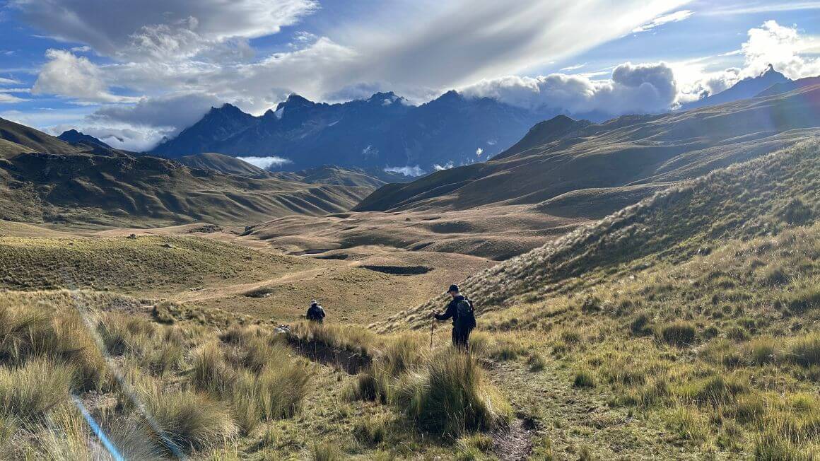 Two people hiking through a valley with massive mountains in the distance and beautiful clouds