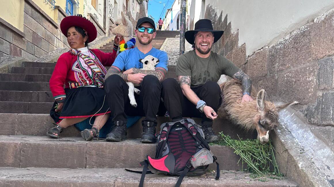 Will and his brother Alex sitting on some steps with a local woman and her alpacas/ llamas in Peru