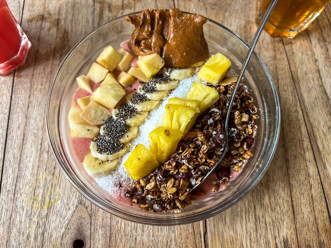 a large light pink smoothie bowl with bananas granola pineapples and peanut butter in a clear glass bowl on top of a wooden table