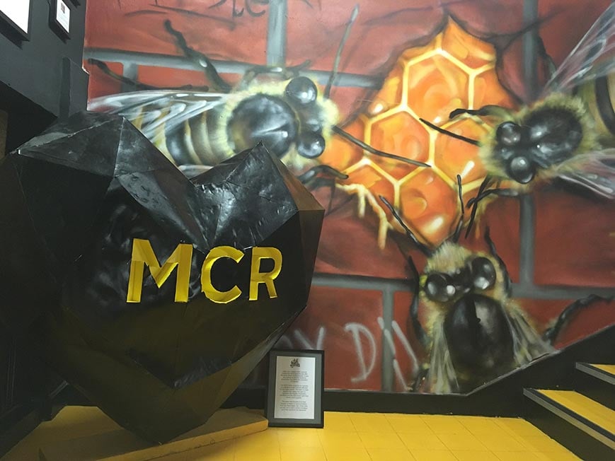 A black heart sculpture with "MCR" engraved in yellow and a mural of some bees behind in Manchester, England, United Kingdom.