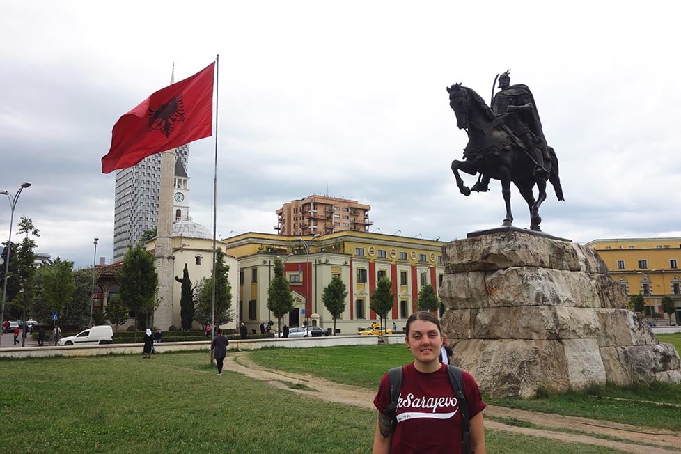 Nic standing in the main square in Tirana, Albania with a massive statue and Albania flag behind them