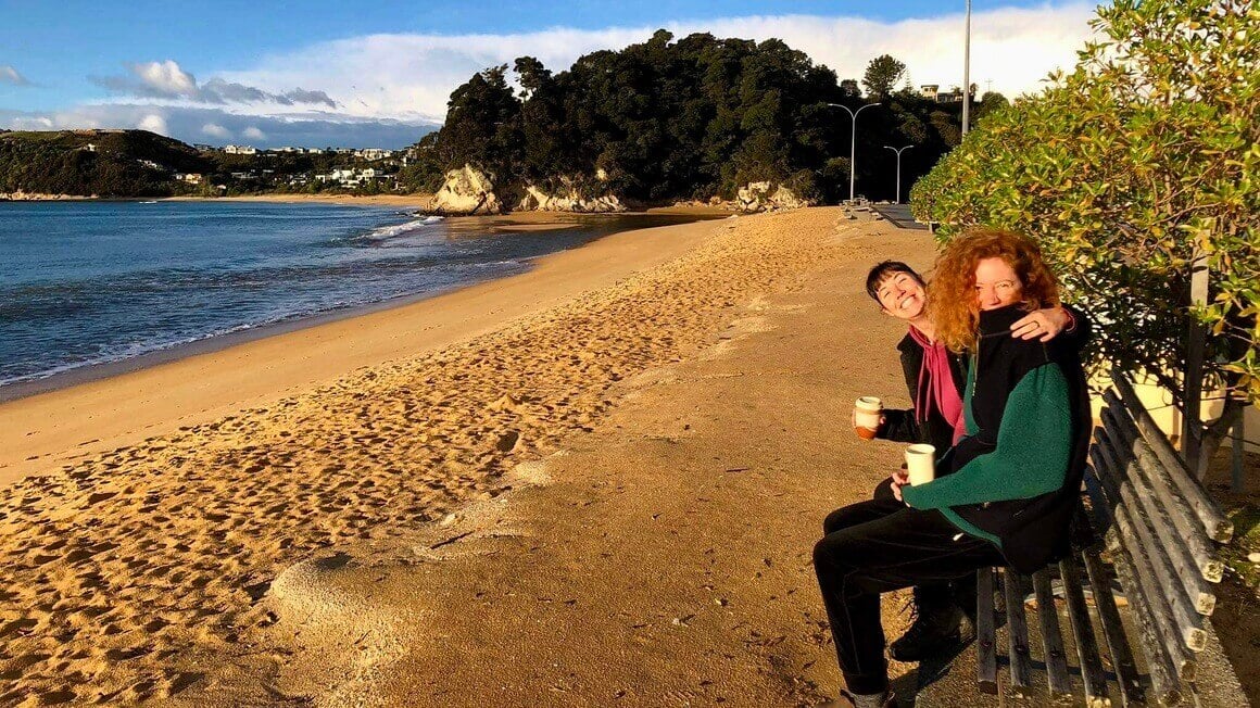 Two girls sitting on a park bench at the beach with their coffee in Kaiteretere, Nelson, Golden Bay, South Island New Zealand