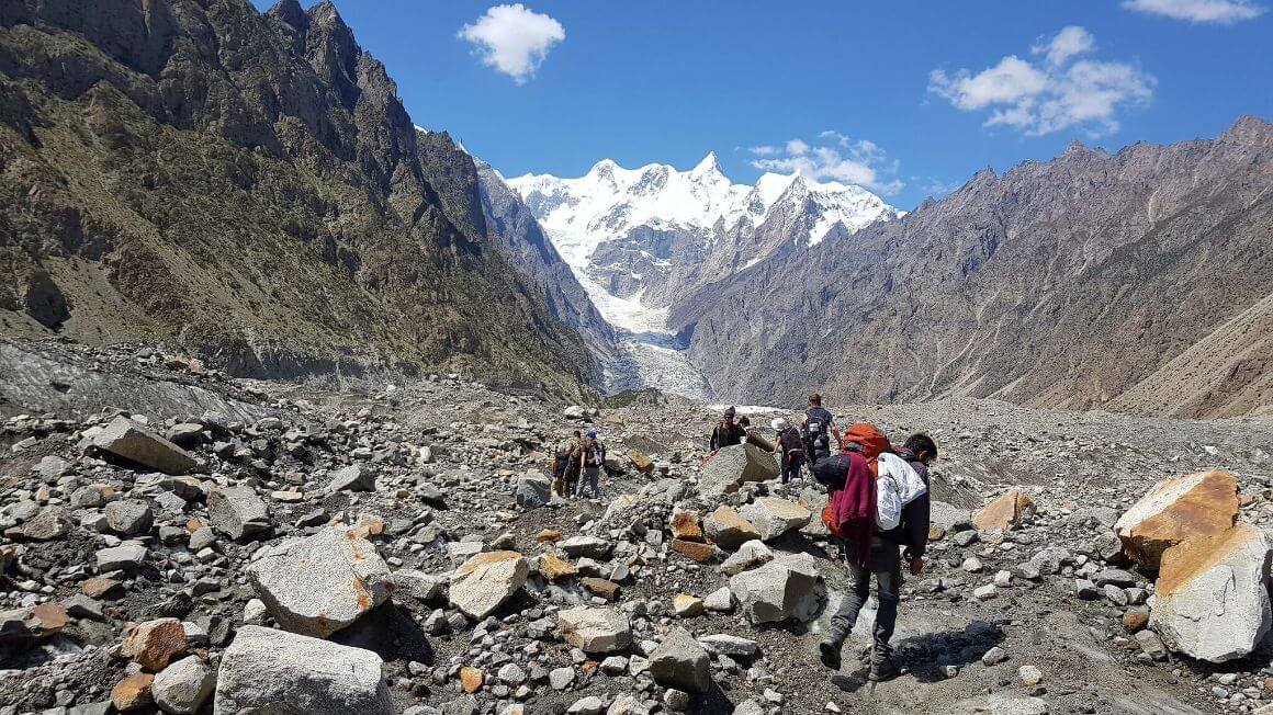 A small group of people walking through a rocky valley with massive mountains on either side and massive snow covered peaks in the distance