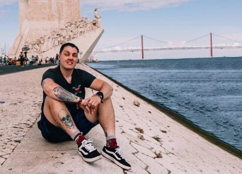 A person sitting along the banks of the river Tagus in Lisbon, Portugal with the monument to the discoveries in the background and the 25 de Abril Bridge.