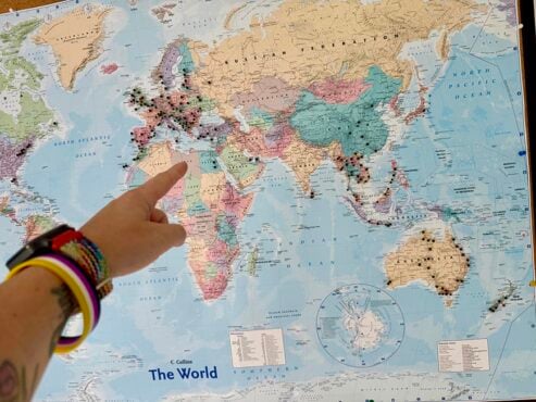 A hand pointing up towards a world map with pins stick in it.