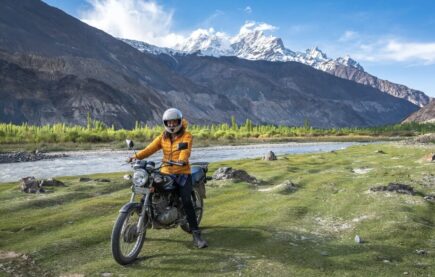 girl in a yellow jacket riding a motorcycle in a lush green meadow with snowcapped peaks in the background